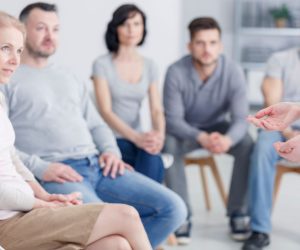 A group of adults sit in chairs, attentively listening to a person speaking in a bright, indoor setting.