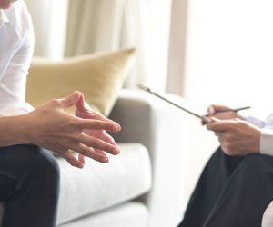 Two people sit on a couch; one gestures with their hands while the other holds a clipboard and pen, suggesting a consultation.