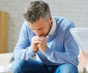 A man sits with his hands clasped, looking down, while another person holds a clipboard and takes notes nearby.