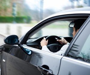 Person driving a black car on a city street, viewed from the passenger side with window partially rolled down.