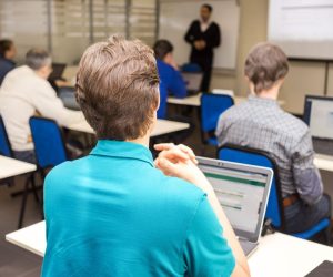 People sit at desks with laptops in a classroom, listening to an instructor standing at the front near a projector screen.