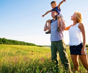 A man carries a child on his shoulders while standing beside a woman in a grassy field under a clear sky.