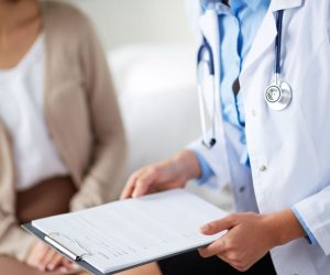 A doctor with a stethoscope reviews a medical chart while speaking with a seated patient.